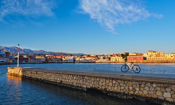 Early morning in Old Venetian harbour of Chania, Crete, Greece. Pier and bicycle in foreground. Chania waterfront promenade and Cretan mountains in background. Golden hour. Bicycle tours, cycling, sightseeing rides. Healthy lifestyle, morning rides