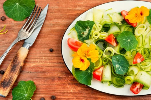Rural salad with vegetables, nasturtium and dressing on wooden table with fork and knife.