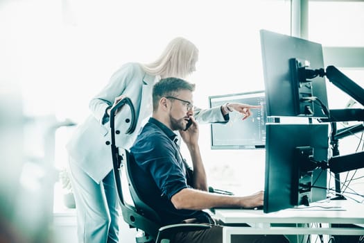 Two thoughtful business colleagues looking at data on multiple computer screens, solving business issue at business meeting in modern corporate office. Business success concept.
