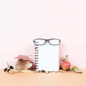 Empty notebook with eyeglasses, an apple and a hedgehog, autumn template with colorful leaves, office desk, pastel color