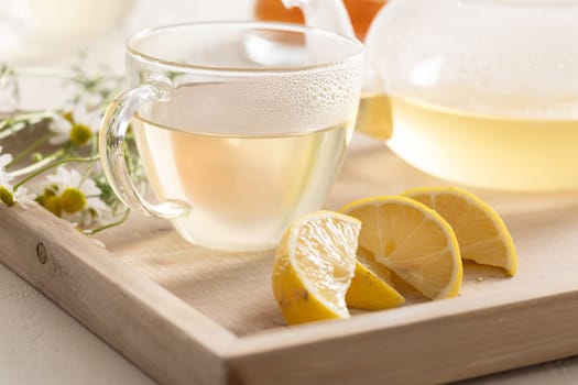 Hot chamomile tea in a glass teacup, sitting on a wooden tray with chamomile flowers and lemon.