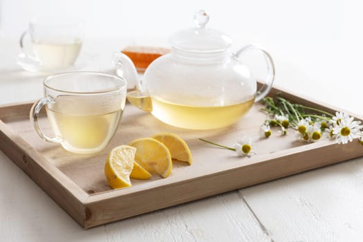 Hot chamomile tea in a glass teapot and teacup, sitting on a wooden tray with chamomile flowers and lemon.