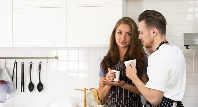 Happy sweet young couple drinking coffee in kitchen together, holding mugs, talking, and enjoying morning leisure time with hot beverage at home.