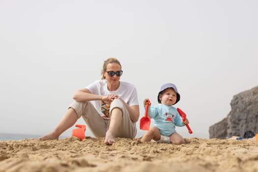 Mother playing his infant baby boy son on sandy beach enjoying summer vacationson on Lanzarote island, Spain. Family travel and vacations concept