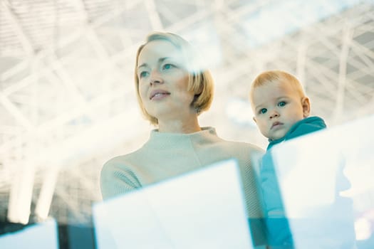 Thoughtful young mother looking trough window holding his infant baby boy child while waiting to board an airplane at airport terminal departure gates. Travel with baby concept