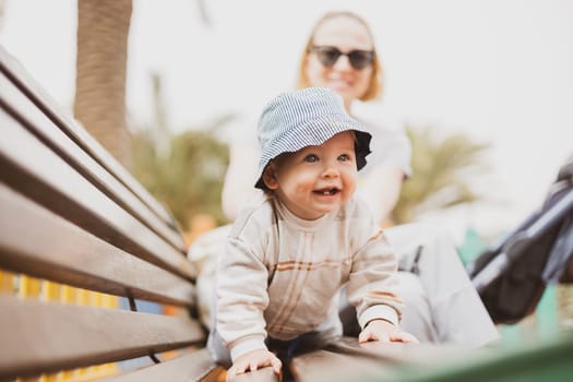 Young mother with her cute infant baby boy child on bench on urban children's playground on warm summer day