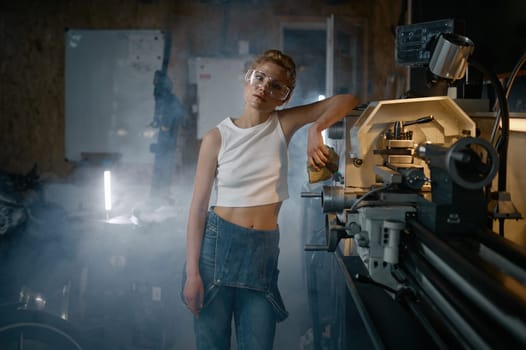 Young woman wearing denim overalls and protective safety goggles after working on lathe turning machine at workshop