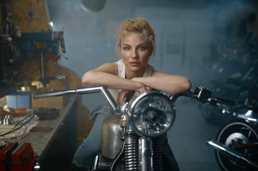 Portrait of beautiful woman biker sitting on motorcycle in garage. Pretty female mechanic posing for camera at workshop