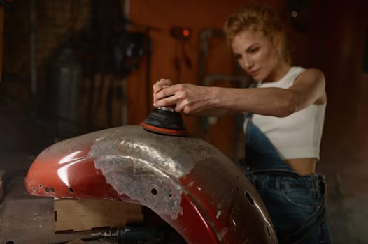 Young woman doing hard job at motorcycle garage using grinder to repair some metal bike part