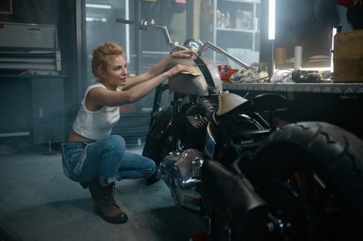 Young pretty satisfied woman mechanic polishing motorcycle fuel tank after repair at workshop