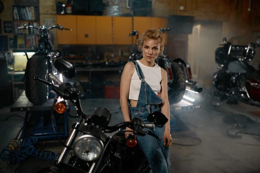 Young woman in denim overalls looking at camera posing at motorcycle garage workshop. Small business concept