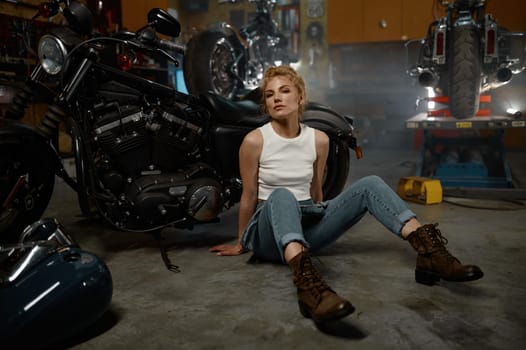 Pretty trendy girl mechanic holding wrench spanner sitting on garage floor nearby motorcycle and looking at camera