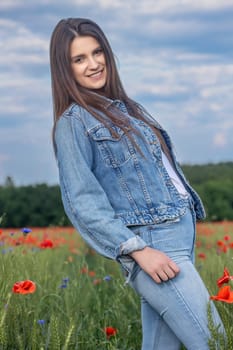 Smiling long haired  brunette girl dressed in blue jeans is posing in red poppies meadow. Vertically.