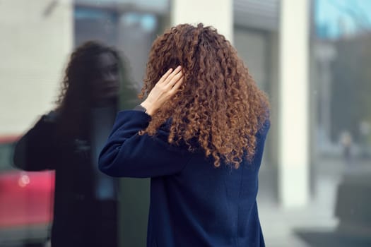 Unrecognizable woman in coat looks at her reflection in the window while being outdoor