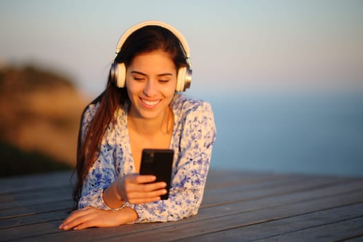 Front view portrait of a happy woman at sunset listening audiobook lying on a wooden terrace on the beach