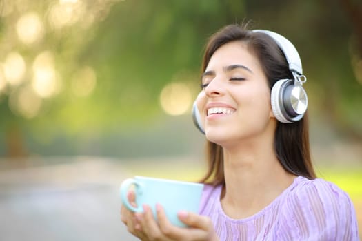 Happy woman wearing headphone listening to music holding coffee mug in a park and breathing fresh air