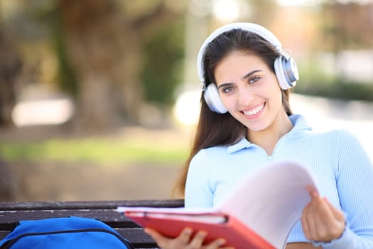 Happy student posing looking at you holding notebook and wearing headphone in a park or campus