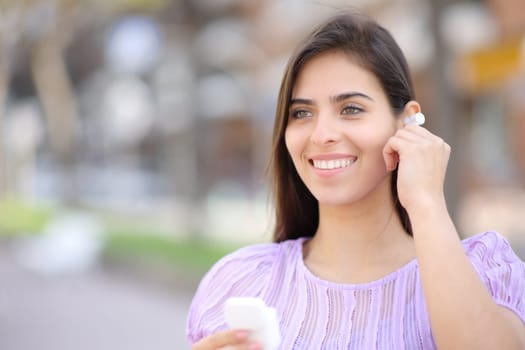 Happy woman putting earbud standing in the street looking away