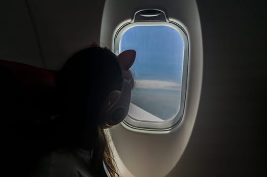 Child girl observing the sky from the window of an aircarft. She has headphones on her head.