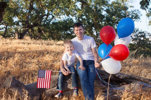 handsome father holding colorful balloons and his little son holding american flag celebrating 4th of July
