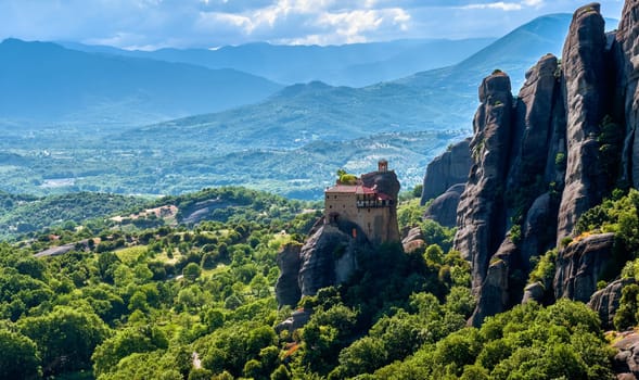 View of monastery of St Nicholaos Anapafsas on top of sedimentary pillar overlooking green hills of Meteora valley at midday. Greece, UNESCO World Heritage.
