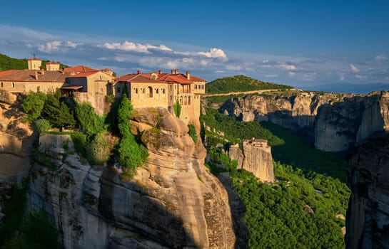 Clifftop Varlaam monastery lit by evening sunlight in Meteora, Greece in typical Meteora landscape of rocky pillars and slopes covered by rich spring foliage. Moni Agias Varvaras Roussanou nunnery in background. UNESCO World Heritage.