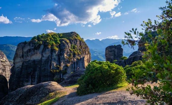 Evening view of typical Meteora landscape, Greece. Massive sedimentary rocks, single cliffs, distant mountains and valley in-between. Blue sky and great clouds. Selective focus. Bush in foreground.