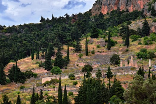 View of famous ancient Greek temple complex known as Delphi oracle. Ruins of temple of Apollo and theater. Tourists rush up and down mountain slopes and paths among pines and cedars.