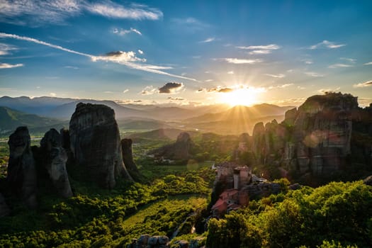 Great view of Meteora, Greece, valley at sunset. Sun set behind distant mountains, sun rays fall on valley, cliffs and rocks, clifftop Moni Agias Varvaras Roussanou nunnery, blue sky and beautiful clouds. Natural lens flares. UNESCO World Heritage.