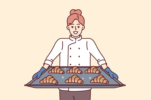 Woman baker holds tray of croissants preparing fresh pastries for sale in own bakery. Girl cook and owner of bakery with smile demonstrates delicious sweet buns for breakfast or lunch