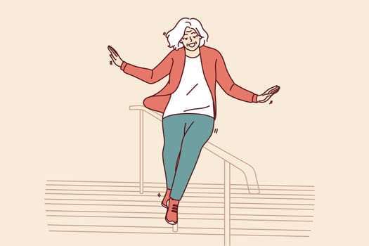 Elderly woman slides down stair railing having fun and being active after retirement. Cheerful elderly woman in casual clothes feels full of energy and laughs regardless of mature age