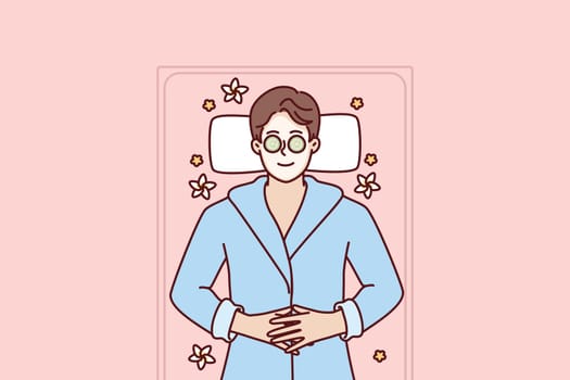 Man with clay mask on face lies in SPA during rejuvenation and anti-wrinkle treatment. Guy with cucumbers on eyes smiles being in SPA salon and restoring strength after hard day work