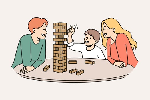 Happy family plays jenga with child enjoying board game and developing fine motor skills in baby. Satisfied boy plays jenga with parents pulling wooden block out of unstable tower.