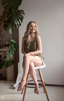 graceful blonde girl in thin dress sits on chair in minimalistic interior. girl has long light hair. she smiles. behind her light wall next to monstera. green dress