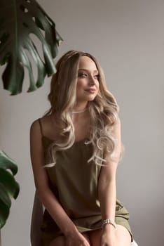 close-up of graceful blonde girl in thin dress sits on chair in minimalistic interior. girl has long light hair. she looks away thoughtfully. behind her light wall next to monstera. green dress