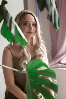 close-up of graceful blonde girl in thin dress sits on chair in minimalistic interior. girl has long light hair. she looks at camera through green monstera leaves. light wall behind her. green dress