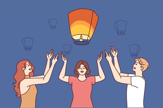 Friends launch chinese air lanterns together to make wish during festive night festival. Man and two women raise hands up standing near flying chinese lanterns illuminating evening sky
