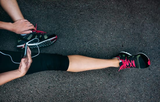 Fit athlete woman runner in sportswear relaxing after training with smartphone. Top view of female lags in sport shoes and outfit on city trail road. Workout lifestyle.