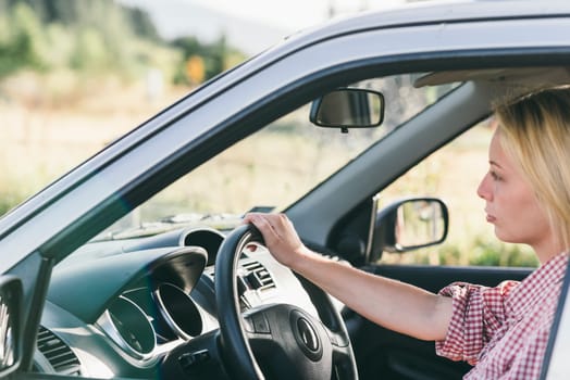 Young caucasian blond woman traveling by car and having fun on her summer holiday journey. Happy girl driving car on countryside road. Roadtrip vacation.
