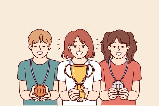 Children with trophies for winning school olympiads or competitions rejoice in awards for leadership positions. Girl with winner cup stands near classmates with medals out for winning competition