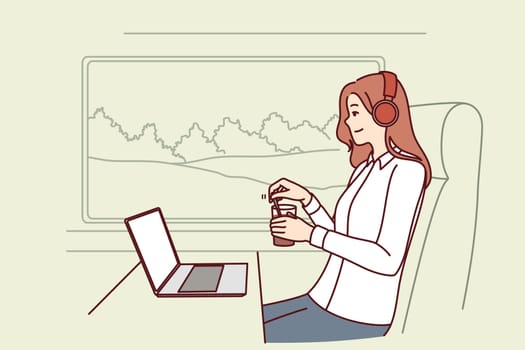 Woman train passenger uses laptop working on trip and drinks coffee sitting near large window in train carriage. Successful girl traveler in headphones sits in train watching movies