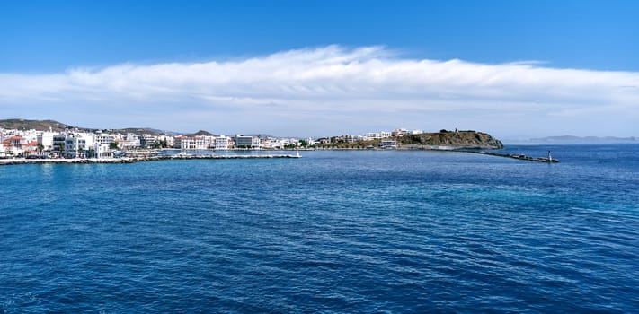 Panoramic sea view of main town of Tinos island and its harbor, Greece. Seascape of small Greek island. White houses, bay, small lighthouse, jetty breakwater. Summer day, great clouds, azure sea. Holidays, sea vacations, Mediterranean island hopping.