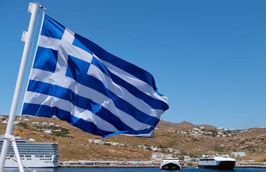 Greek flag waving against typical Greek island landscape. Cruise liners and ferries moored in background. Bright summer day. Island hopping, adventures and exploring Mediterranean sea. Selective focus.