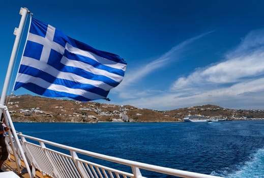 Greek flag waving against typical Greek island landscape. Cruise liners and ferries moored in background. Bright summer day. Island hopping, adventures and exploring Mediterranean sea. Selective focus.