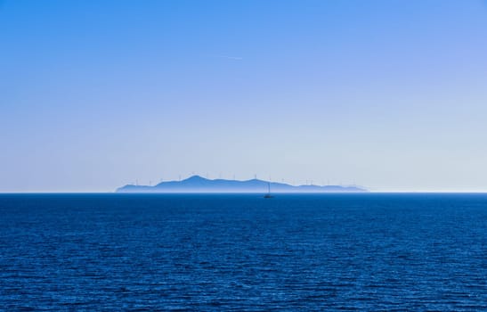 Seascape of calm azure sea and blue sky with no clouds. Small sailing boat and distant island in hazy background dotted with silhouettes of electric turbine windmills. Mediterranean sea, Greece.