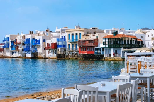 Little Venice of Mykonos main town or Chora, Cyclades, Greece. Romantic neighborhood with bars, cafes, restaurants in whitewashed old fisherman houses hanging on cliffs above sea waves. Colorful tables and chairs by shoreline. Summer day, blue sky