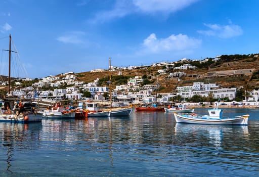 Beautiful summer day sunshine in typical marina of Greek island. Fishing boats, yachts moored at jetty. Whitewashed houses on hills and by waterfront. Mediterranean lifestyle, vacations. White fishing boat in foreground. Mykonos, Cyclades, Greece.