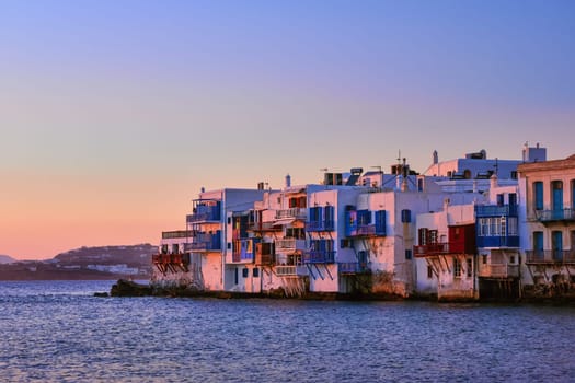 Beautiful sunset view of Little Venice in Mykonos, Greece. Romantic neighborhood with whitewashed bars, cafes, restaurants, old fisherman houses hanging on cliffs above sea waves.