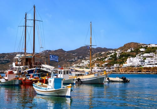Beautiful summer day and sunshine in typical marina of Greek island. Colorful fishing boats and tourist cruise yachts moored at jetty. Whitewashed houses on hills and by waterfront. Mediterranean lifestyle, vacations. Mykonos, Cyclades, Greece.