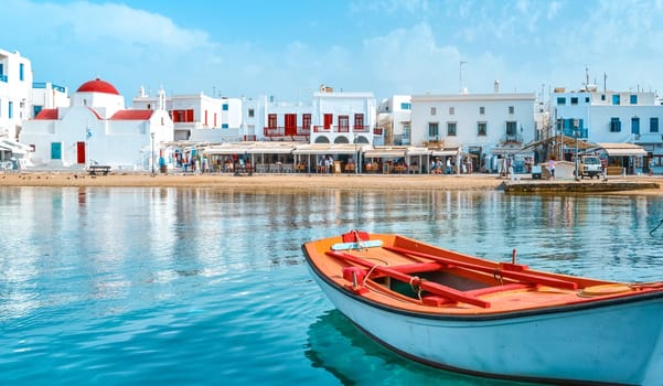 Beautiful summer day sunshine on Greek island. Fishing boats moored at jetty. Whitewashed houses by waterfront. Red dome church, restaurants, shops, fish market stalls. Mediterranean lifestyle. Small boat in foreground. Mykonos, Cyclades, Greece.